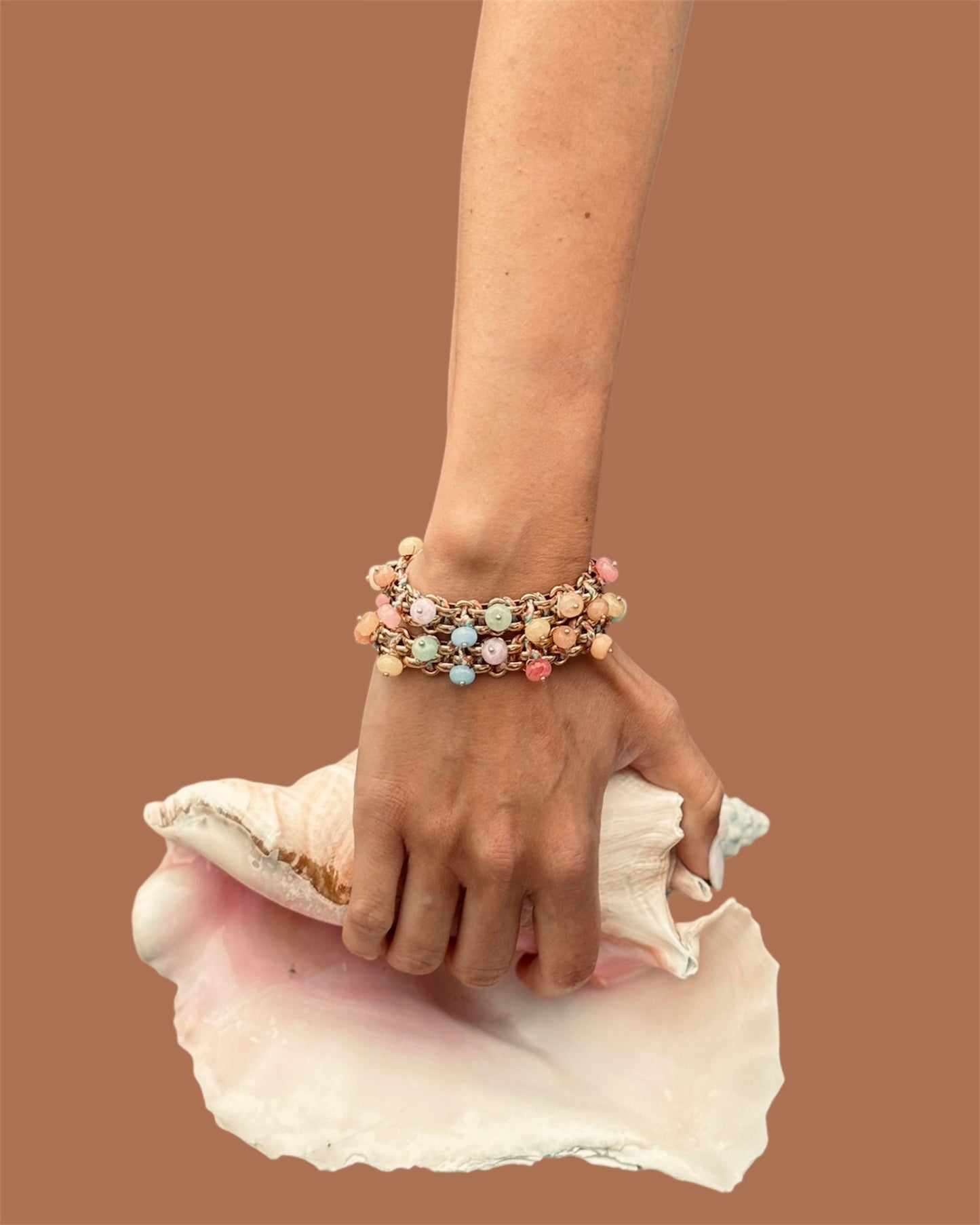 Bracelet Sugar Divaine porté au poignet, empilé en accumulation, avec perles pastel sur chaîne dorée et cordon tressé, posé sur un coquillage.