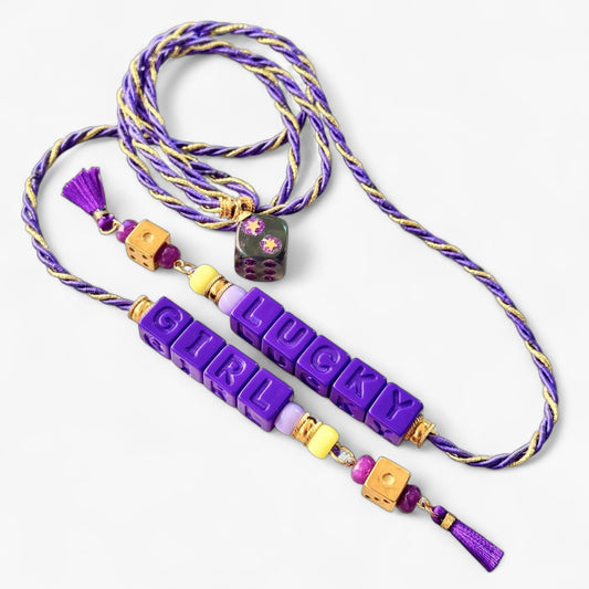 Collier Casino Club violet et doré avec lettrage “LUCKY GIRL”, perles jaunes et violettes, dés orange et pompons violets.
