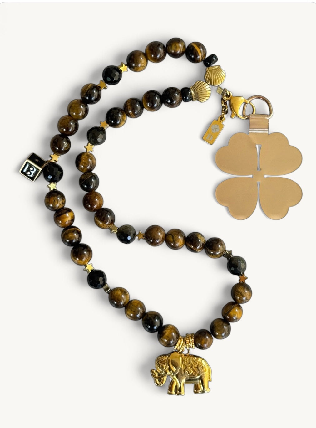 Lanière de téléphone DIVAINE Lucky Jungle en perles d’œil de tigre et acier inoxydable, ornée d’un pendentif éléphant doré et de charms porte-bonheur, sur fond blanc minimaliste.